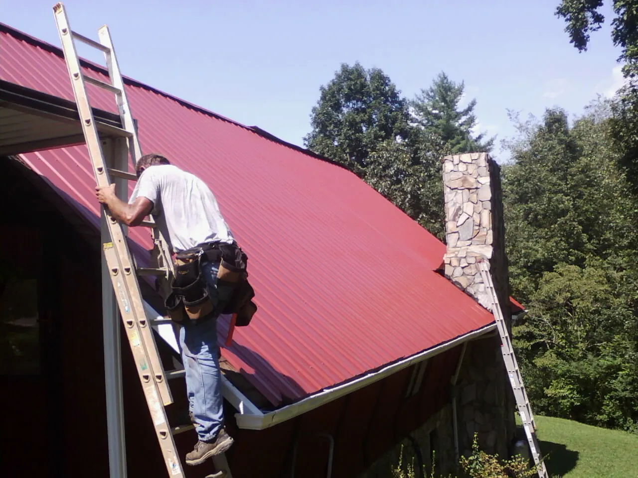 Roofer on a ladder beside a red metal roof and stone chimney — Soffit & Fascia Repair by Roofing Brothers in Buckley
